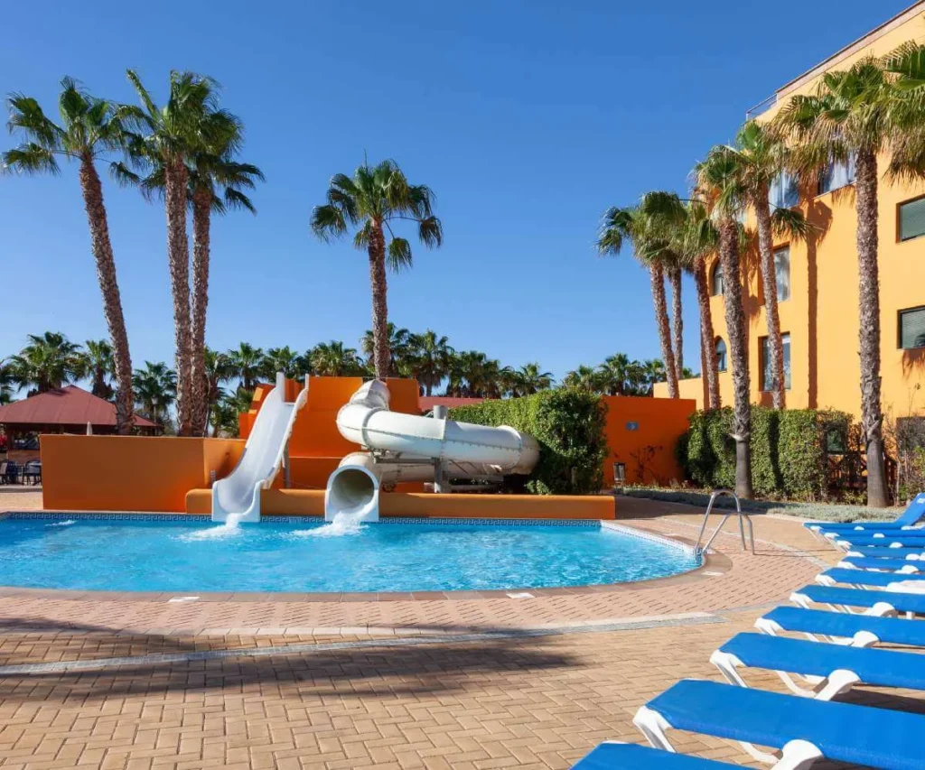 Piscina de hotel con tobog&aacute;n, tumbonas azules y palmeras al cielo despejado