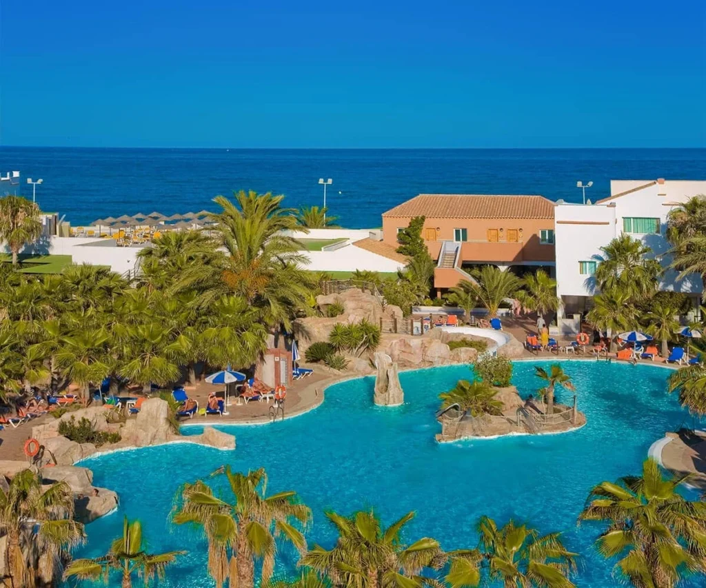 Piscina azul con palmeras, caba&ntilde;as junto al mar y sombrillas azules