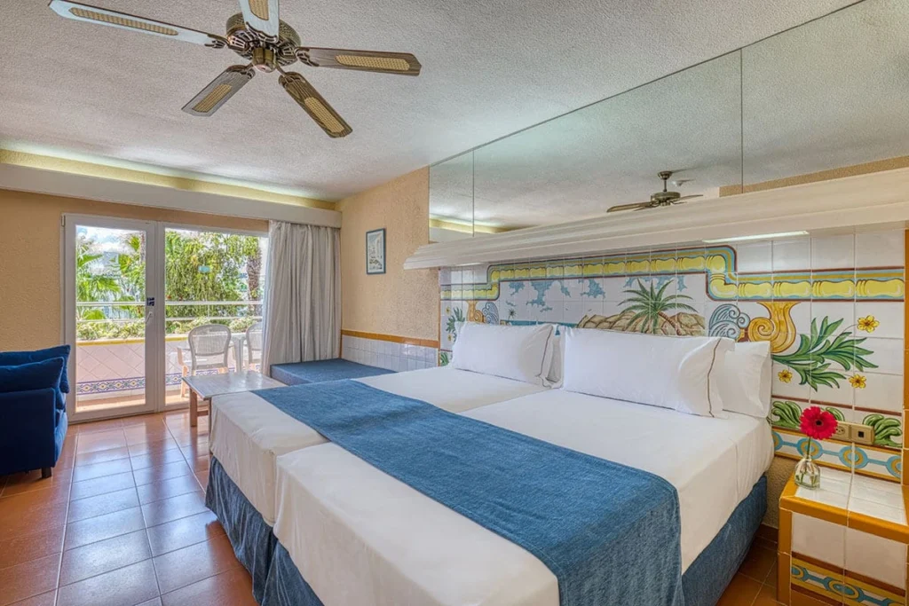 Dormitorio luminoso con dos camas, cojines azules, ventilador y salida a terraza.