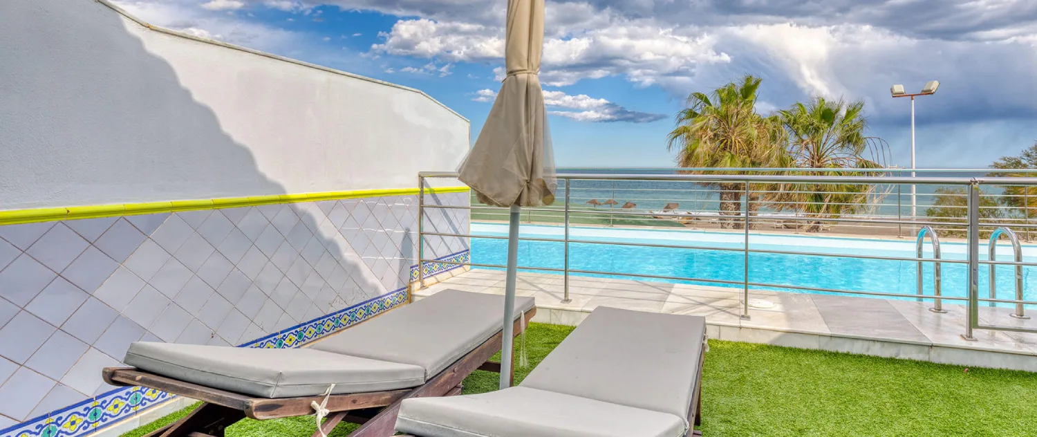 Terraza con c&eacute;sped, tumbonas y piscina frente al mar, bajo cielo nublado