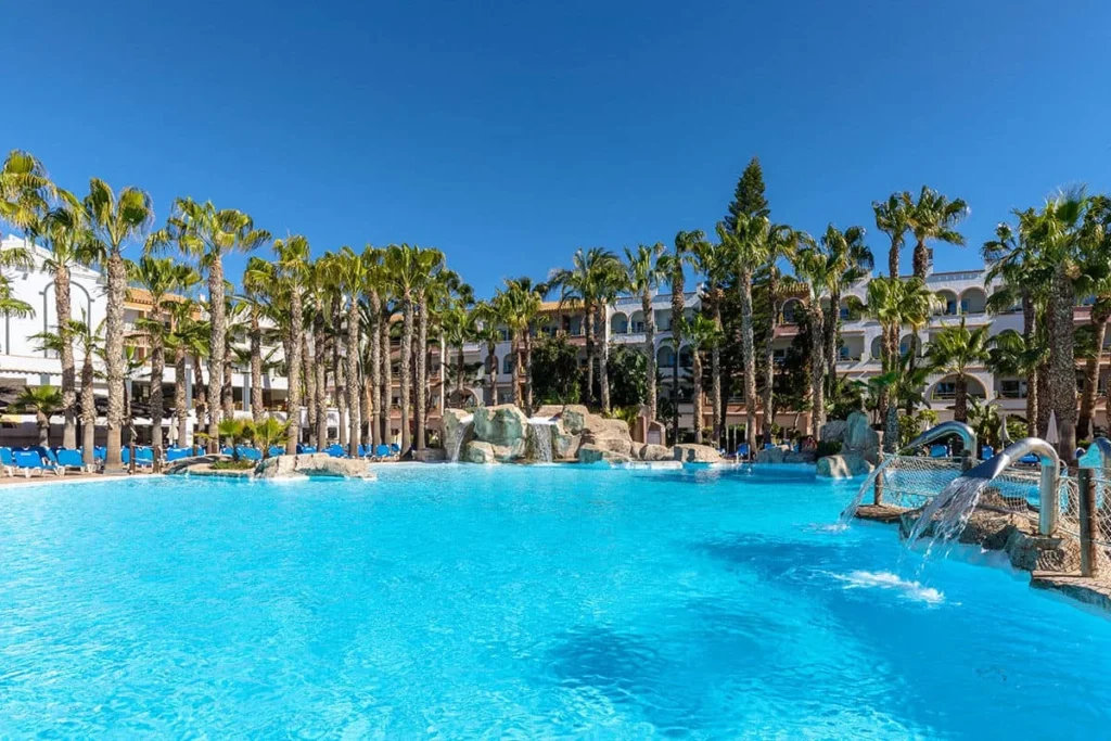 Piscina azul con palmeras y tobog&aacute;n, junto a un hotel al sol