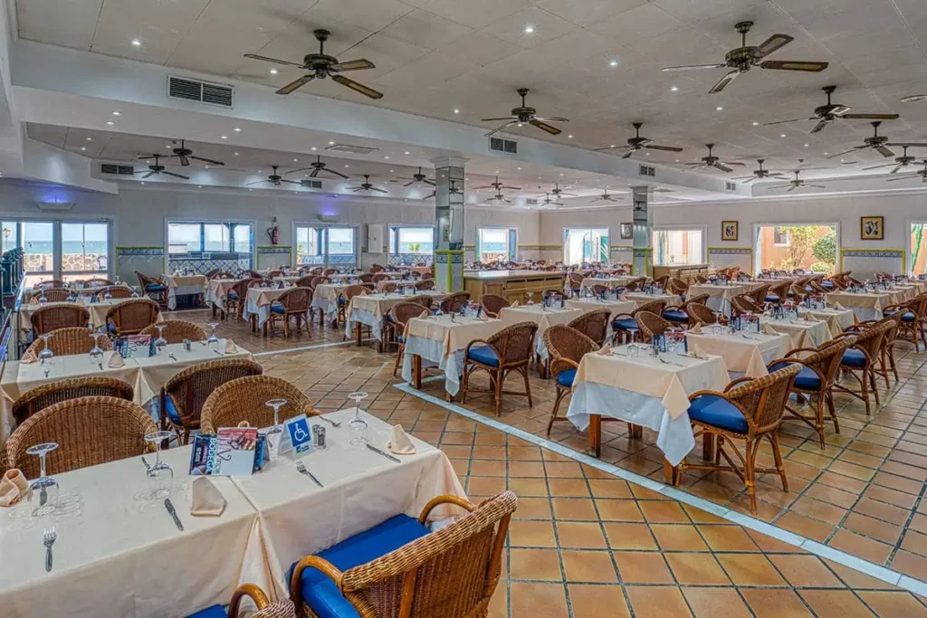 Comedor de hotel con mesas cubiertas, sillas de mimbre y ventiladores de techo