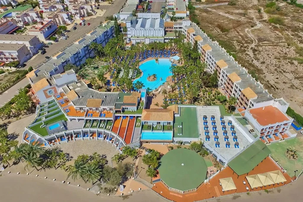 Vista a&eacute;rea de complejo hotelero con piscinas, jardines y &aacute;reas deportivas junto a playa