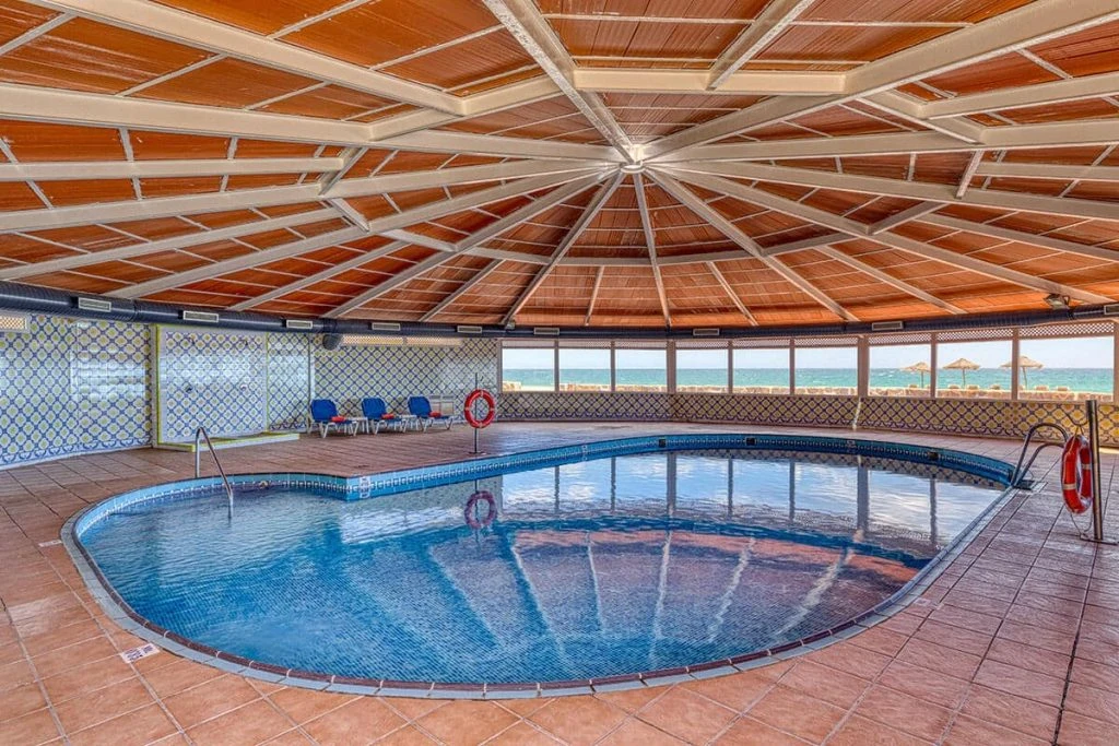 Piscina cubierta con techo de madera y vista al mar, luces y tumbonas