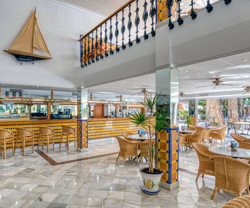 Interior de hotel con barandal azul, mesas y sillones de mimbre y palmera