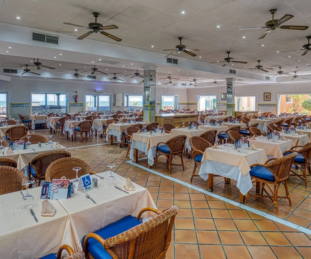Comedor de restaurante con mesas servidas y sillas de mimbre, l&aacute;mparas de techo
