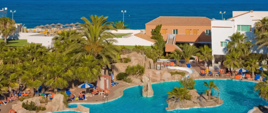 Piscina de hotel junto al mar, palmeras, tumbonas y sombrillas azules