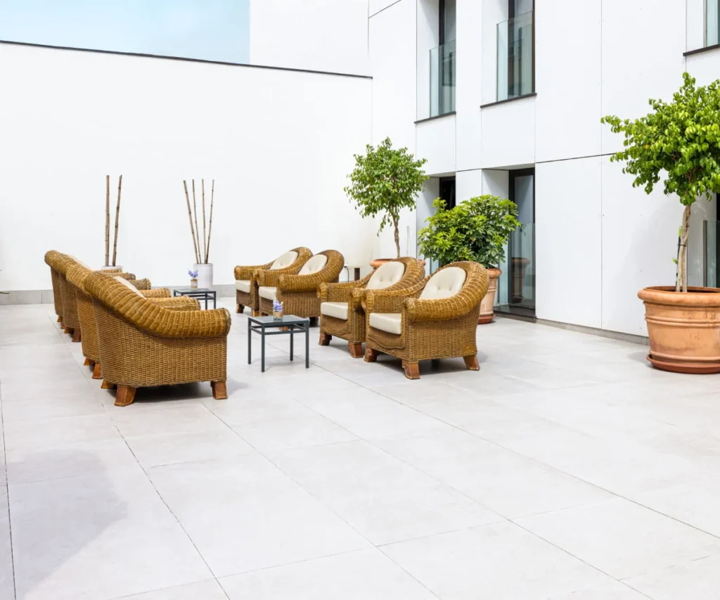 Patio moderno con sillones de rat&aacute;n, mesas y plantas junto a un edificio blanco