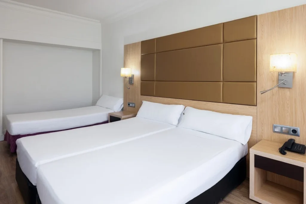 Habitaci&oacute;n de hotel moderna con dos camas, cabecero acolchado y l&aacute;mparas.