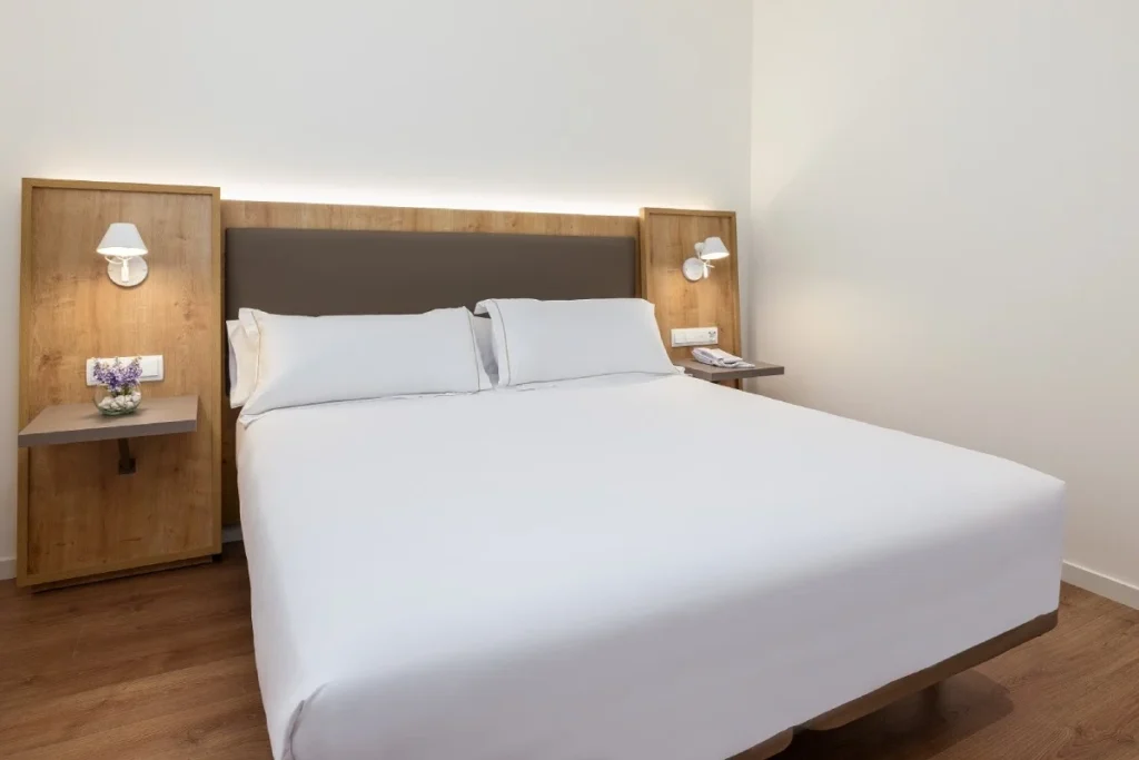 Habitaci&oacute;n con cama doble blanca, mesillas de madera y l&aacute;mparas de pared