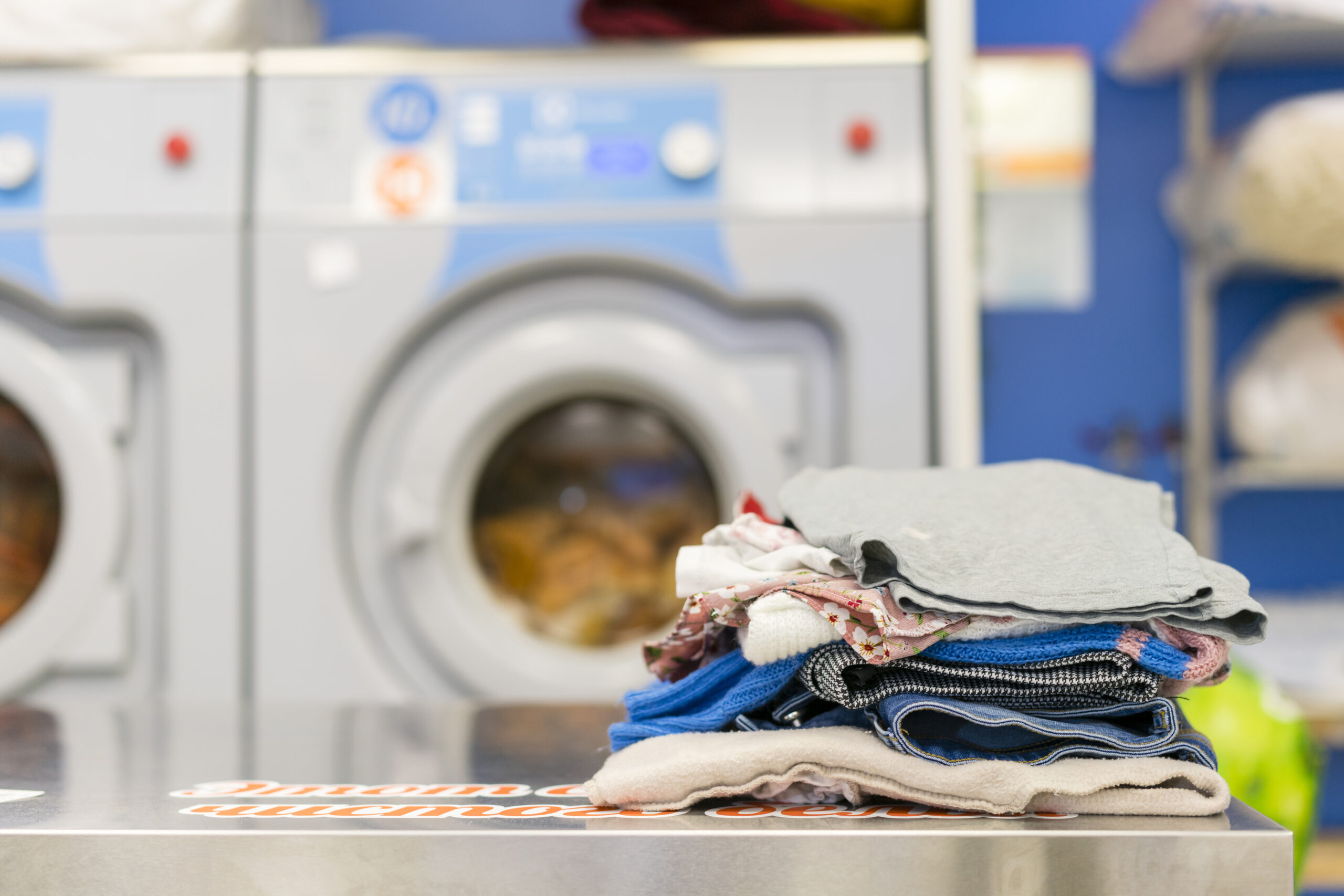 Zona de una lavandería con ropa doblada en primer plano y lavadoras de fondo