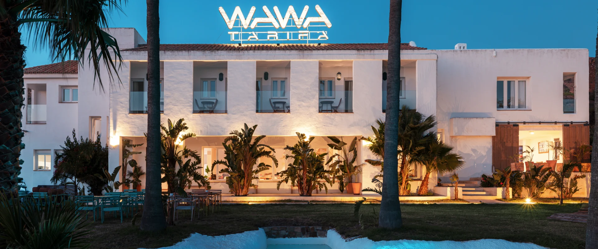 Hotel con fachada blanca, palmeras y logo &ldquo;WAWA&rdquo; iluminado al atardecer