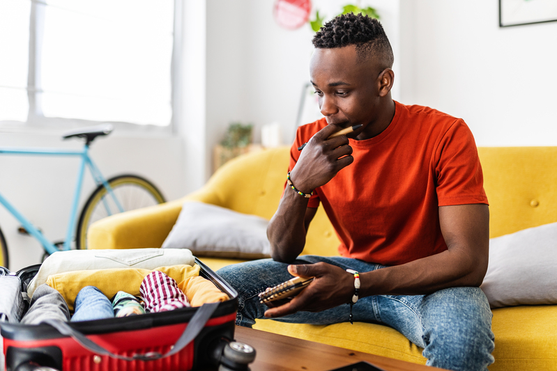 Young adult man packing clothes to the suitcase and writing travel checklist