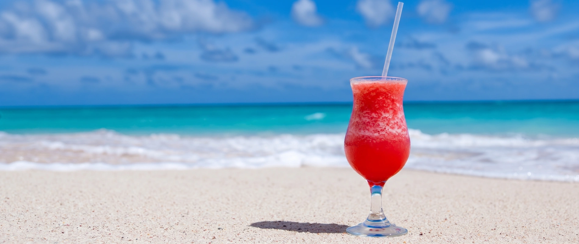 C&oacute;ctel rojo en copa con pajita, en la playa con mar y cielo azul al fondo.