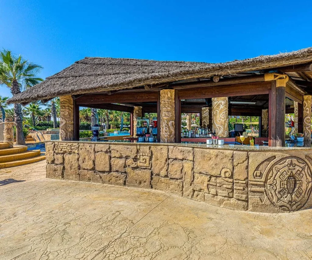 Bar r&uacute;stico de madera con techo de paja y decoraci&oacute;n tallada junto a piscina