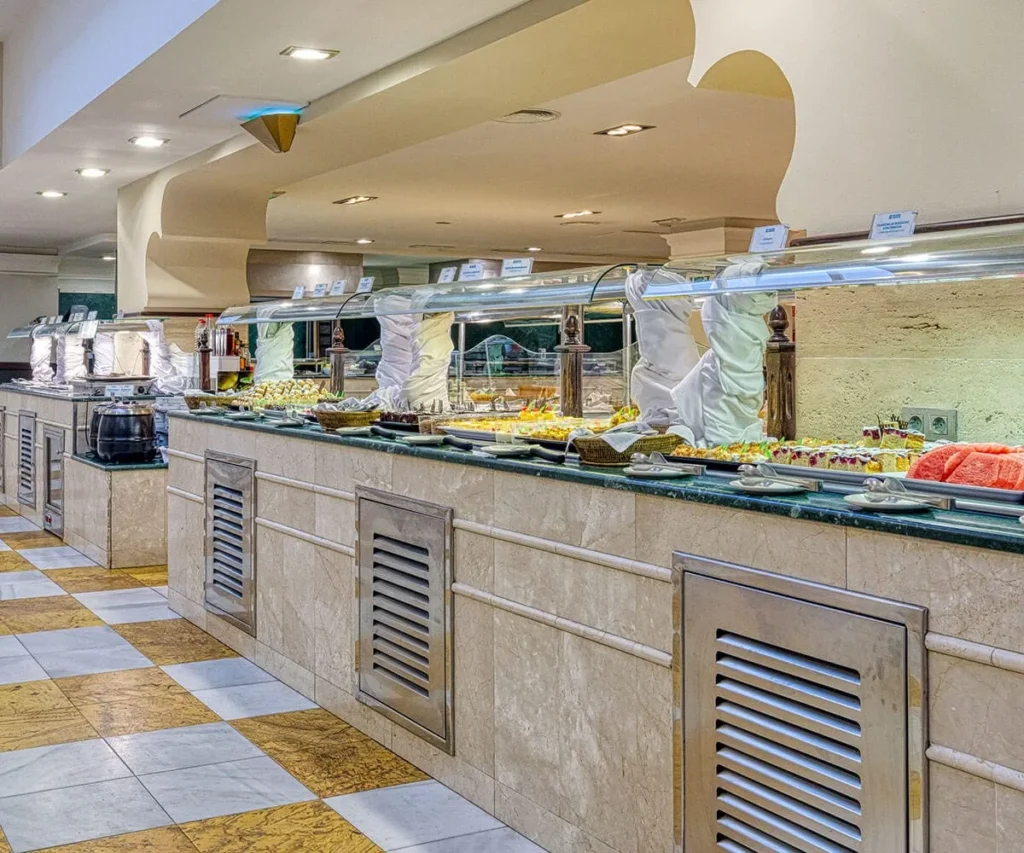 Buffet con mostrador refrigerado, bandejas de comida y personal con guantes