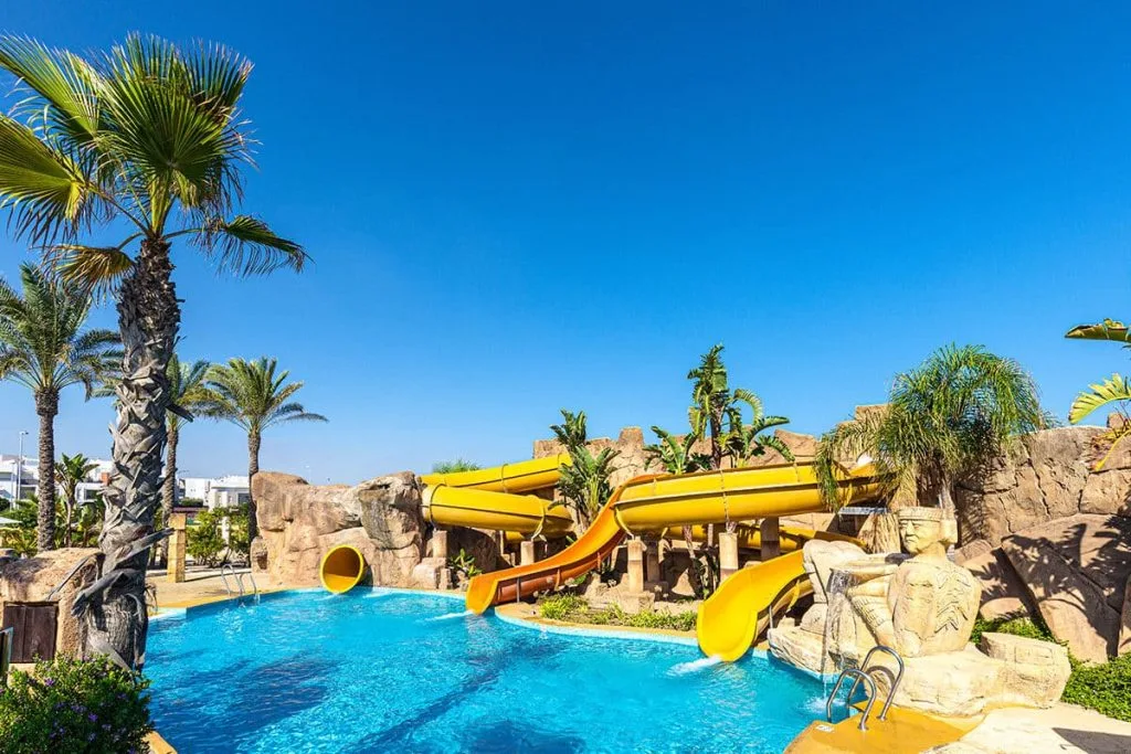 Piscina azul con palmeras y toboganes amarillos junto a rocas, bajo cielo azul.