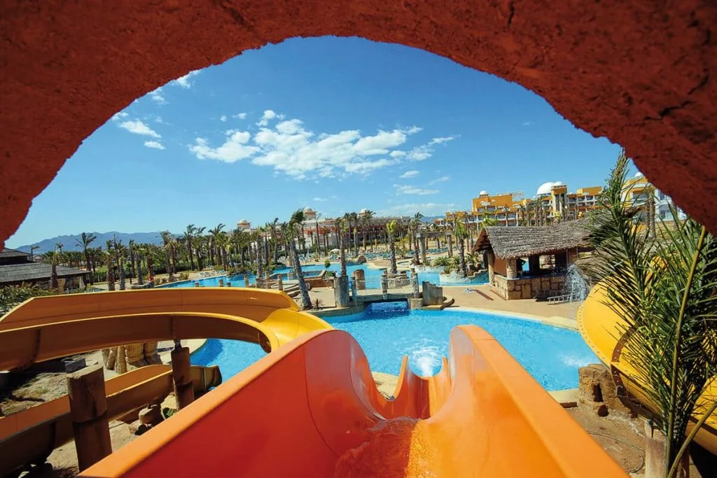 Piscina tropical con tobog&aacute;n naranja y palmeras, vista al cielo azul.