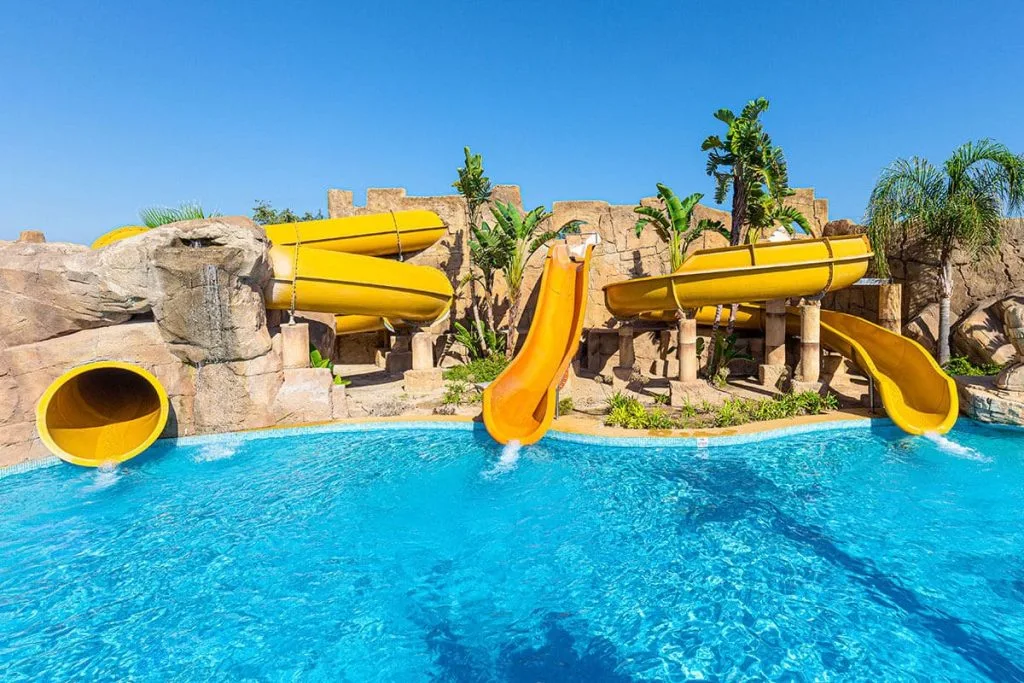 Piscina azul con toboganes amarillos y fondo de palmeras y rocas.