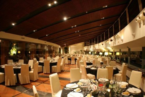 Restaurante elegante con techo curvo, mesas redondas preparadas y cubiertas de tela blanca. Ambiente cálido e iluminado.