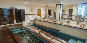 Instalaciones interiores de un circuito de spa con un camino de guijarros y agua para los pies, rodeado de plantas en macetas y tumbonas al fondo.