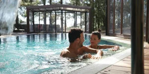 Una pareja joven sonr&iacute;e y conversa dentro de una piscina de agua cristalina, con una cascada en primer plano y un entorno boscoso visible al fondo.