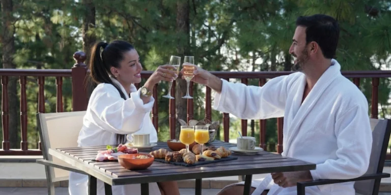 Pareja en albornoces blancos brindando con copas de champán durante el desayuno en terraza con vistas a bosque de pinos, escapada romántica entre semana en Tenerife.