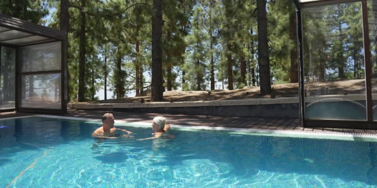 Una pareja disfruta de un ba&ntilde;o en una piscina exterior rodeada de una terraza de madera y un denso bosque de pinos de fondo.