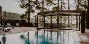 &Aacute;rea de piscina exterior con un tramo semicubierto por una estructura de cristal y aluminio. El agua de la piscina refleja los pinos altos de un bosque circundante. La zona est&aacute; rodeada por un suelo de parqu&eacute; de madera, tumbonas alineadas con sombrillas cerradas y un muro de piedra natural al fondo.