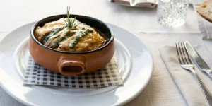 Presentaci&oacute;n de un plato culinario en el Restaurante La Vendimia. En una cazuela de barro marr&oacute;n con asas, colocada sobre un plato blanco con una servilleta de cuadros blancos y negros, se sirve un alimento gratinado con queso fundido y decorado con l&iacute;neas de salsa verde y una ramita de romero. A la derecha, se aprecian un tenedor y un cuchillo sobre un mantel claro.