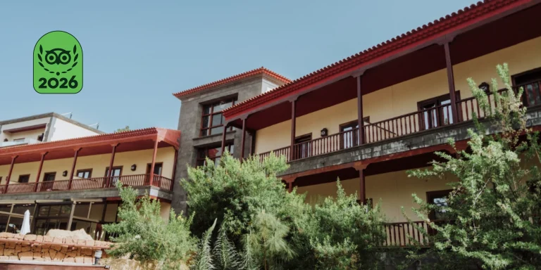 Fachada exterior del Hotel Spa Villalba, de estilo tradicional canario con balcones corridos de madera oscura y paredes de tonos crema y piedra. En primer plano hay &aacute;rboles y arbustos verdes, y en la esquina superior izquierda se muestra superpuesto el logotipo verde de TripAdvisor con el premio "Travellers' Choice 2026".
