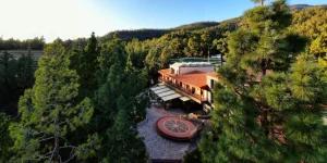 Vista a&eacute;rea del Hotel Spa Villalba integrado en medio de un denso bosque de pinos canarios bajo la luz del atardecer. Se observa el edificio principal con tejado de tejas arcillosas, una terraza con una plaza empedrada en la que destaca un dise&ntilde;o circular conc&eacute;ntrico, y colinas boscosas que se extienden hacia el horizonte.