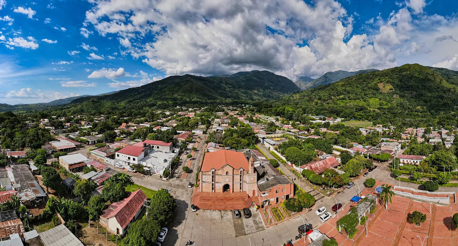Vista a&eacute;rea de un pueblo con una iglesia en el centro, rodeado de monta&ntilde;as y con un cielo parcialmente nublado.