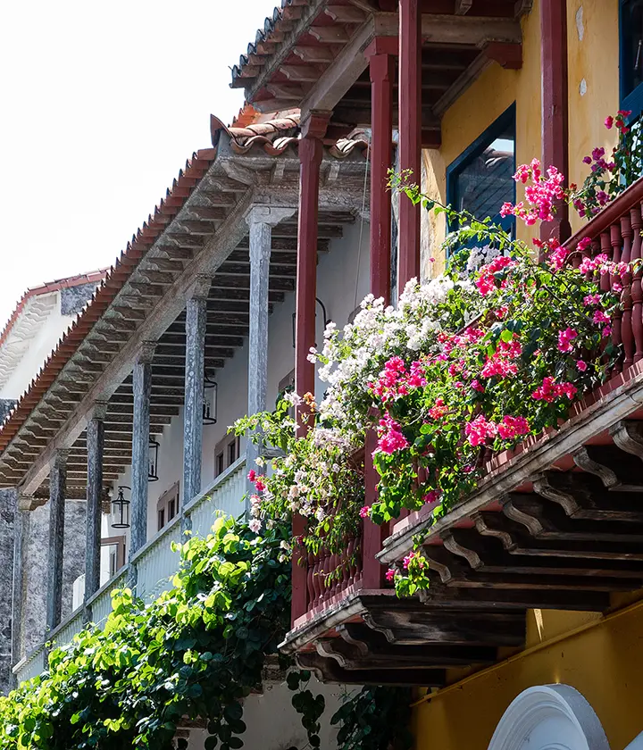 Balc&oacute;n colonial con flores coloridas y enredaderas, bajo un techo de tejas rojas y estructura de madera.