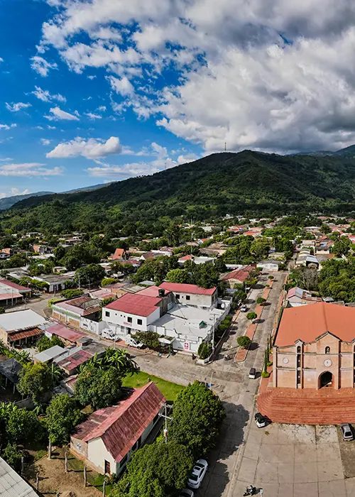 Vista a&eacute;rea de un pueblo rodeado de monta&ntilde;as, con calles, casas y una iglesia bajo un cielo parcialmente nublado.
