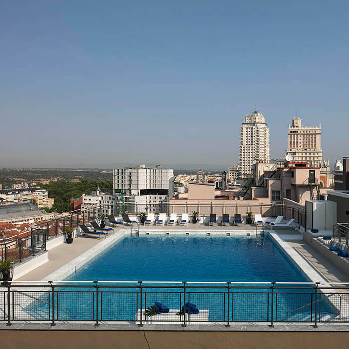Piscina en la azotea con vistas a una ciudad, rodeada de edificios altos y cielo despejado.