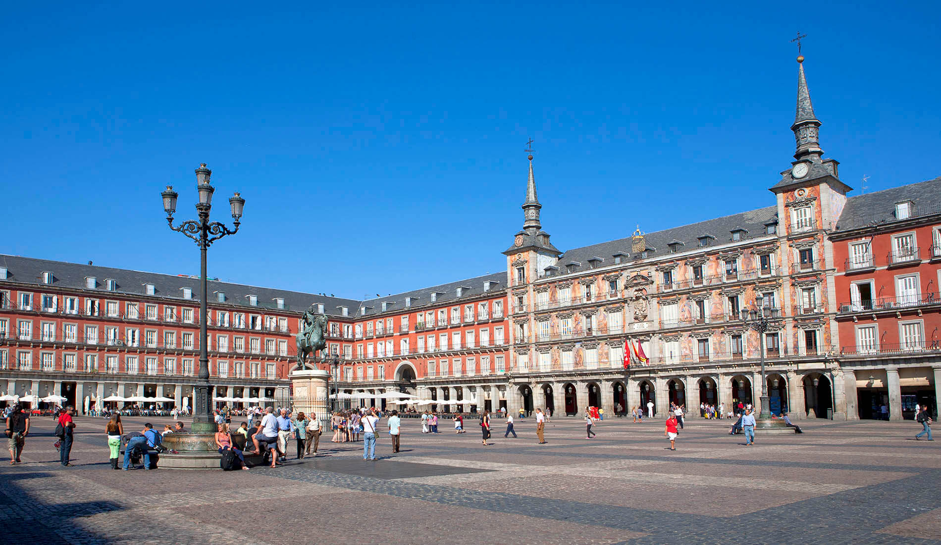 Plaza Mayor de Madrid con visitants, edificios rojos y estatuas bajo un cielo azul claro.