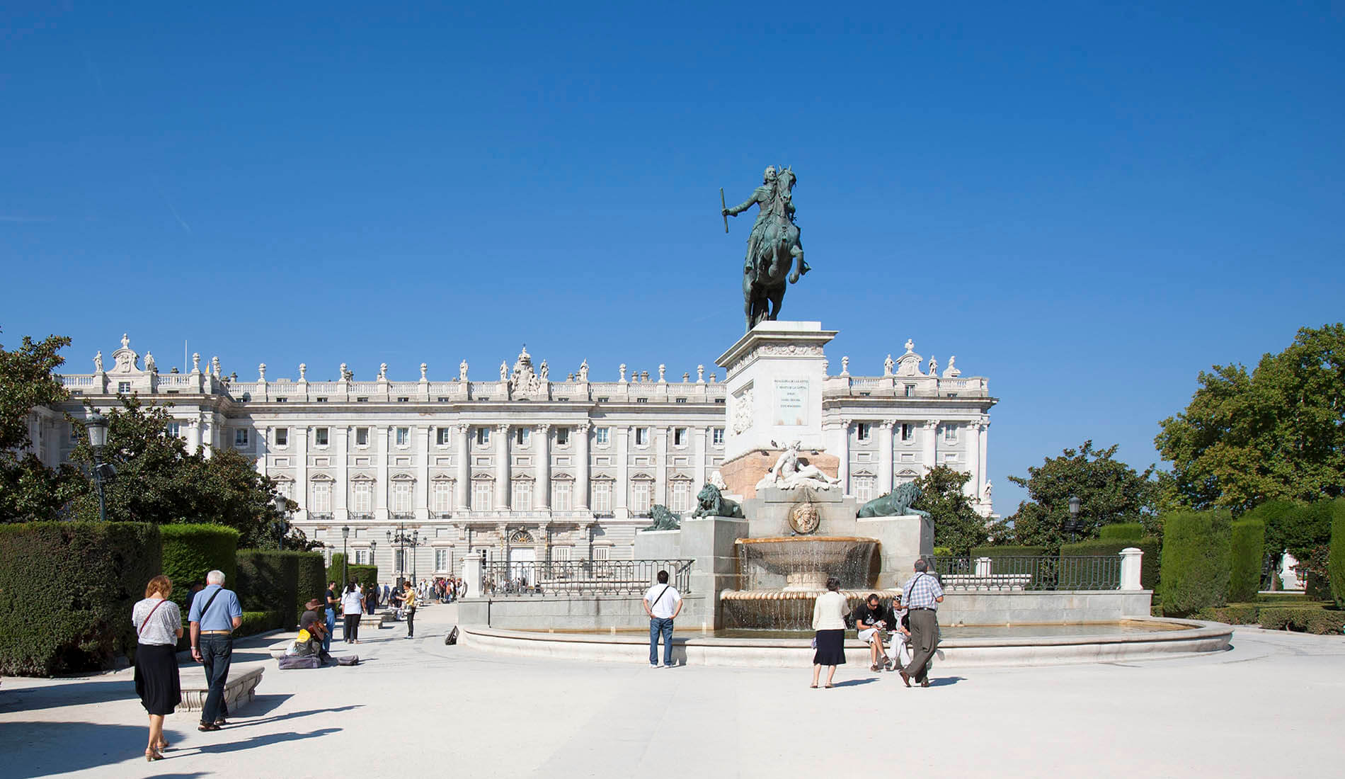 Estatua ecuestre y fuente frente a un palacio con cielo azul y personas paseando.