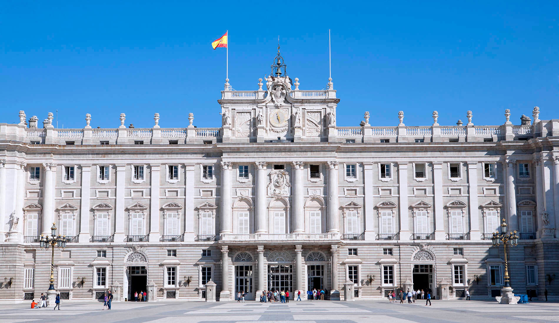 Palacio Real de Madrid con cielo azul y visitantes en la entrada.