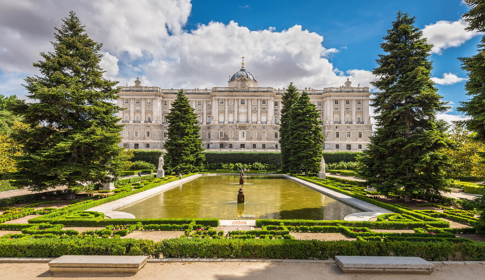 Jardín con estanque frente a un majestuoso edificio clásico, rodeado de árboles y cielo nublado.