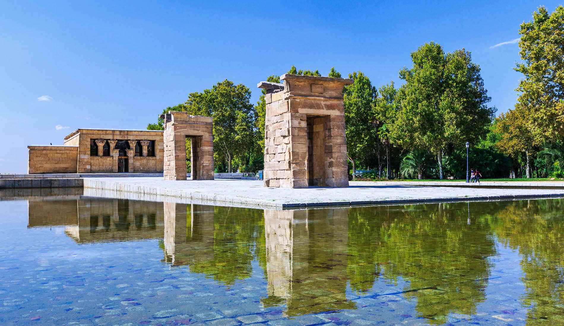 Templo egipcio en un parque, reflejado en un estanque bajo cielo azul.