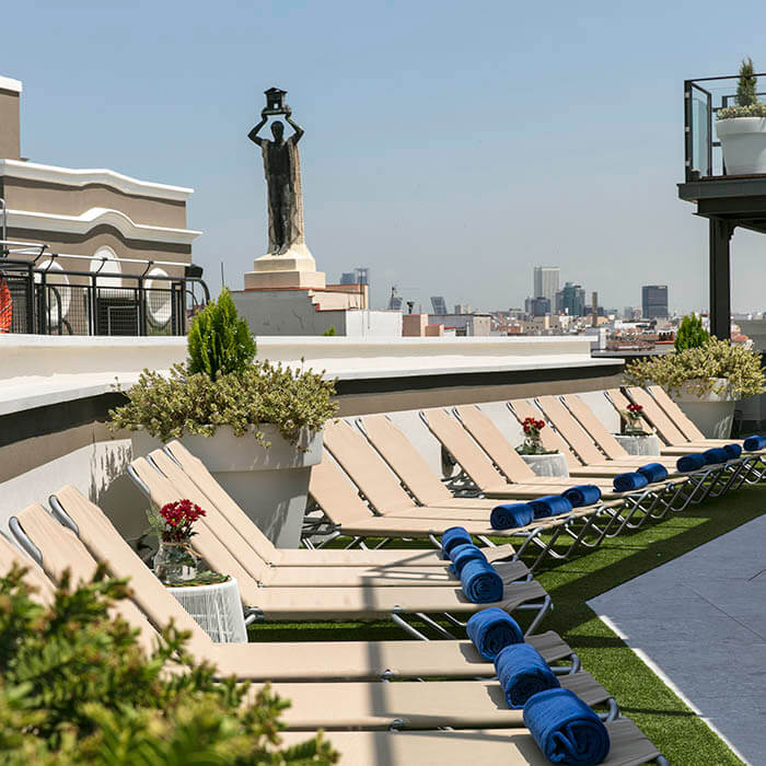 Terraza con tumbonas beige y toallas azules, vista a la ciudad y estatua en el fondo.