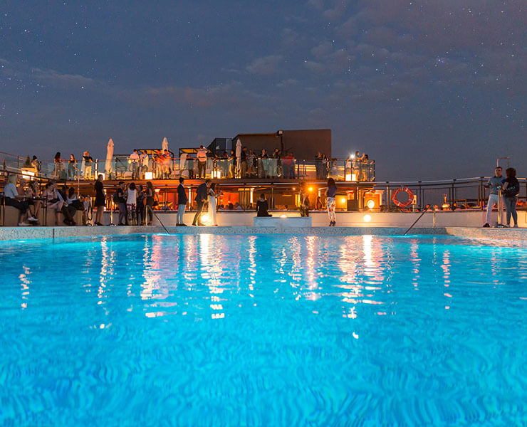 Personas disfrutando de una terraza iluminada junto a una piscina bajo un cielo estrellado.