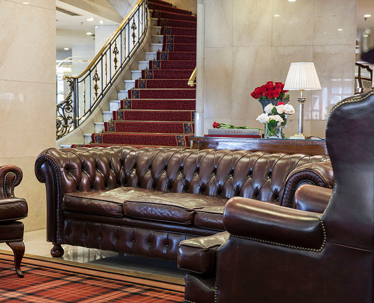 Sofá de cuero marrón en sala elegante, escalera y mesa con lámpara y flores al fondo.