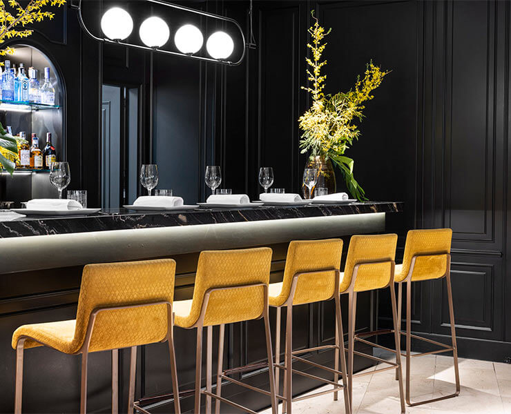 Barra elegante con sillas amarillas y luces modernas; botellas y flores decoran el fondo.