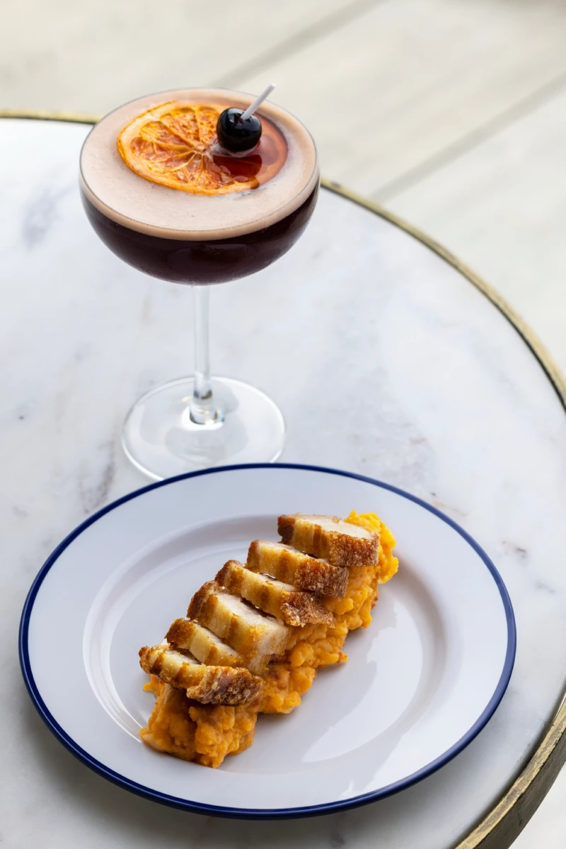 C&oacute;ctel oscuro con rodaja de naranja y cereza, y plato de carne sobre pur&eacute; en mesa de m&aacute;rmol.