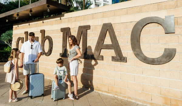 Familia frente a un muro con el texto "club MAC", con maletas y ropa veraniega.